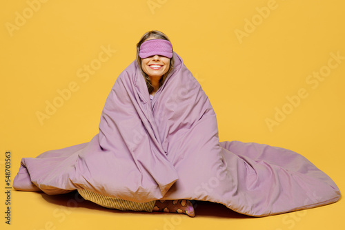 Full body young smiling cheerful happy fun woman she wears purple pyjamas jam sleep eye mask rest relax at home sit wrap covered blanket duvet isolated on plain yellow background. Night nap concept.