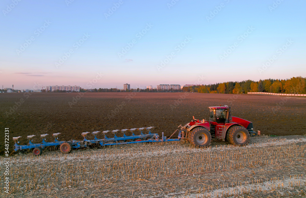Obraz premium Tractor Plowing field on sunset. Red Tractor with Plough on Plowed. Ploughing and Soil Tillage. Agricultural Tractor on Cultivation Field for Sowing Seeds. Big Tractor During Field Cultivating.