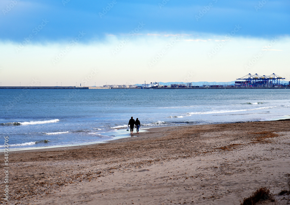 People walk along the coastline of the sea in autumn season. A man and a woman walk barefoot on beach near sea. Persons walking on empty beach in winter season. Sea with beach at seaport.