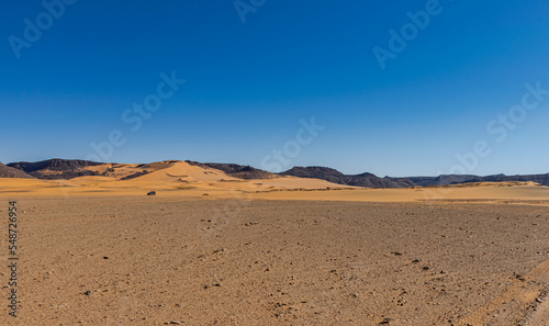Sahara Desert reg stony plain in foreground and sand dune with rocky mountains, 4x4 off-road tiny car far away. Clear blue sky in a sunny day. Tadrart Rouge, Algeria. 