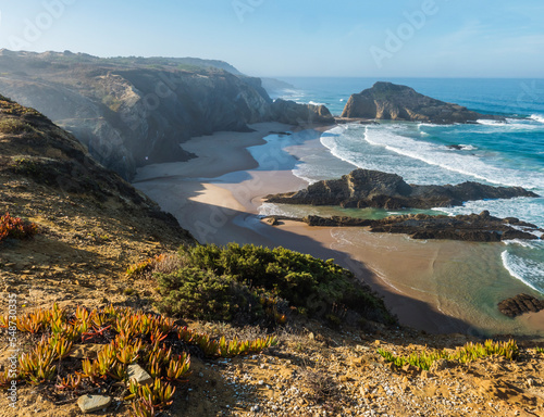 View of empty Praia da Zambujeira do Mar beach with ocean waves, cliffs and stones, wet golden sand and green vegetation at wild Rota Vicentina coast, Odemira, Portugal. photo
