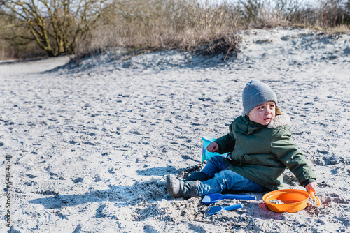 Eager enthusiast kid sits on a beach playing with sand toys in winter time. Fervid or fervent child dressed or covered against the cold weather is sitting on a strand, playing on a sunny, winter day photo