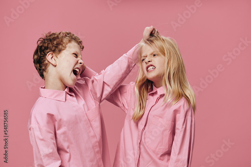 a boy and a girl of school age in pink clothes stand on a pink background and fight fiercely with each other