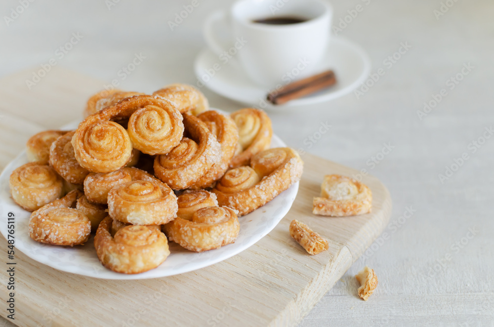 Puff pastry cookies with sugar on a white plate with a small white cup with coffee and a cinnamon stick. The concept of homemade baking. copy space Horizontal orientation. Selective focus Puff pastry cookies with sugar on a white plate with a small white cup with coffee and a cinnamon stick. The concept of homemade baking. copy space Horizontal orientation. Selective focus