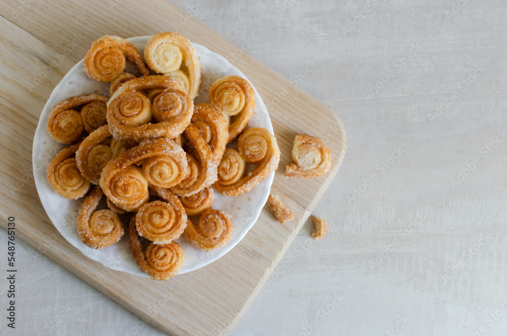 Fototapeta premium Puff pastry cookies with sugar on a white plate on a wooden cutting board. The concept of homemade baking. copy space Horizontal orientation. Top view. Selective focus