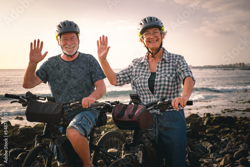 Bye bye. Senior cheerful couple riding on the pebble beach with electric bicycles at sunset. Authentic elderly retired life and sustainable mobility concept