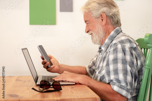 Senior caucasian man sitting at a wooden table in coffee shop using laptop holding his smart phone. Elderly man reading message on cellphone while typing on keyboard