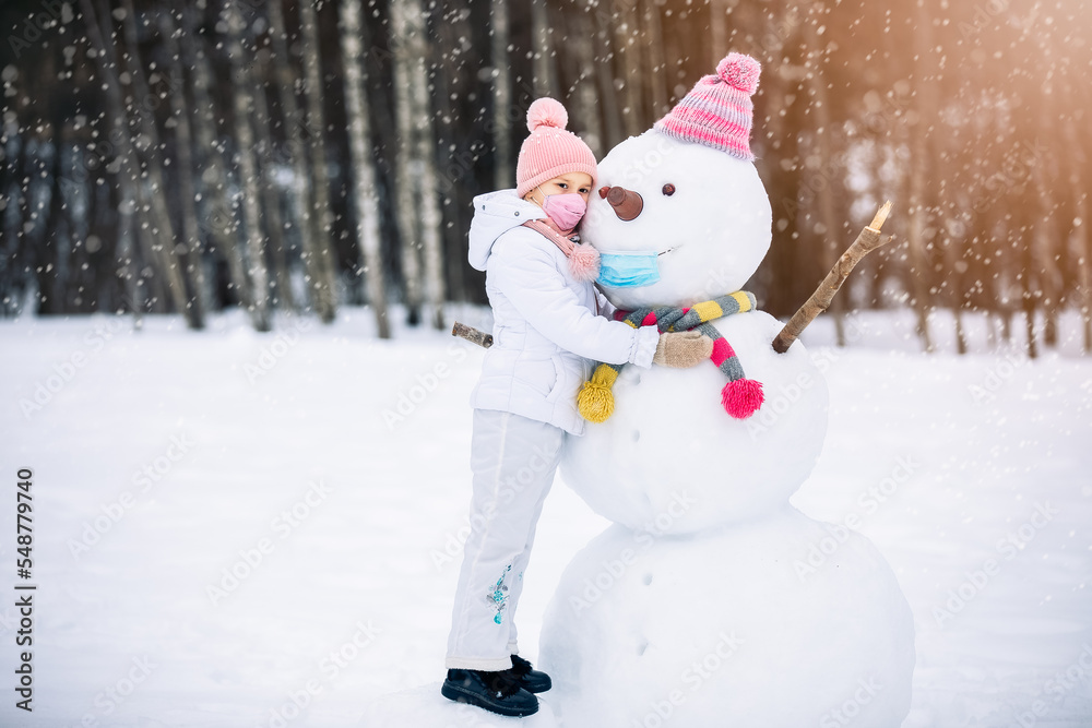 A child girl wearing a mask herself and on a snowman ,playing with a snowman on a winter walk in nature