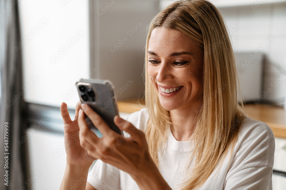 Blonde white woman smiling and using cellphone in kitchen Blonde white woman smiling and using cellphone in kitchen