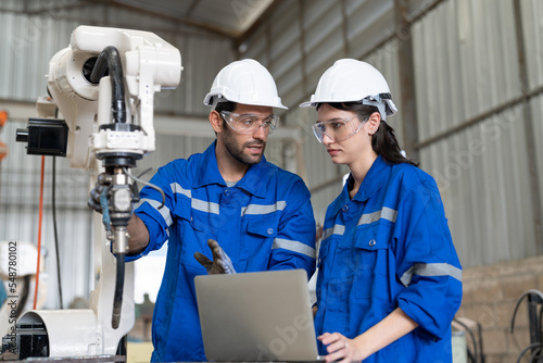 Male and female engineer using laptop working with robot arm welding machine in industrial factory. Team of technician automation robot in uniform explains robot arm system.