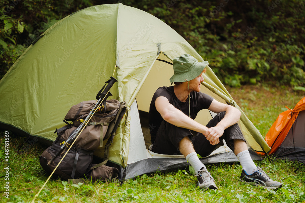 White young man resting in tent while hiking in green forest - obrazy, fototapety, plakaty 