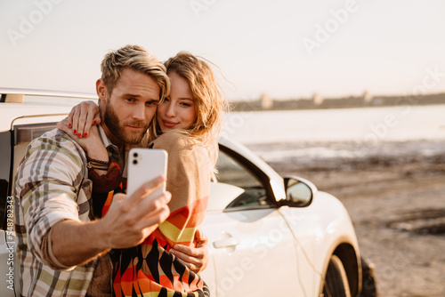 Happy white couple taking selfie photo by car while walking at seashore