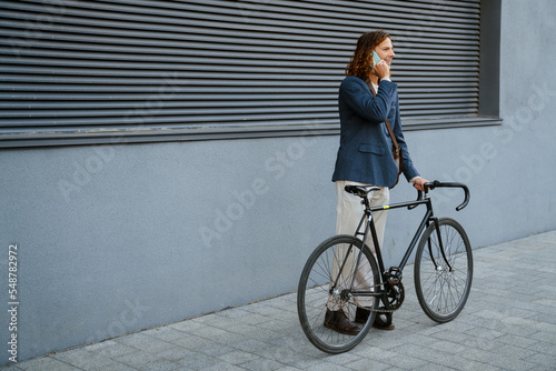 Young joyful man talking on cellphone while standing with bicycle outdoors