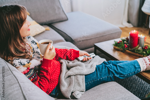 Relaxed woman in plaid and warm socks drinking hot tea with remote controller on sofa at home while watching movie, TV with christmas decoration atmosphere. Cozy and comfortable winter concept. © okrasiuk