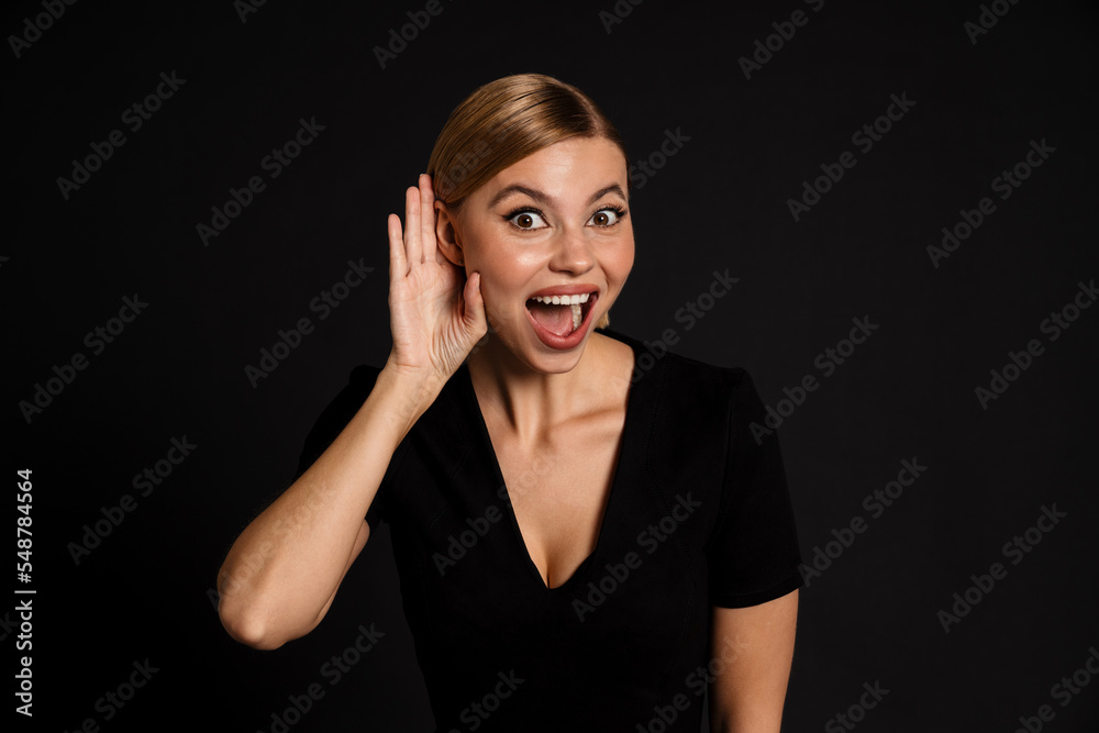Young amazed woman with hand at the ear to hear better standing isolated over black background Young amazed woman with hand at the ear to hear better standing isolated over black background