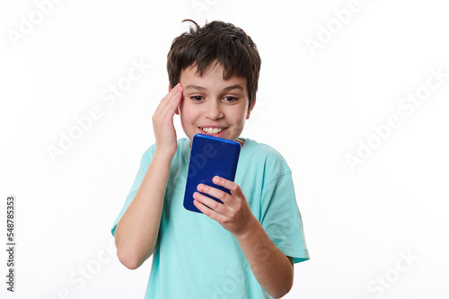 Happy excited teenage school boy holding his hand on his temple, expressing surprise while reading news on his smartphone, isolated over white background with copy space for text