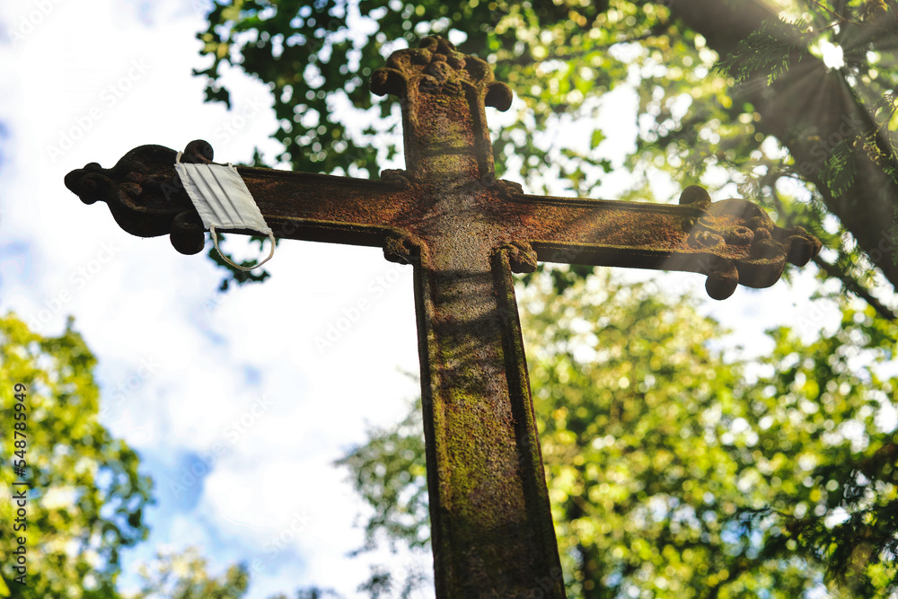 A rusty metal cross with a facemask hanging on it in an old cemetery. Beautiful sun rays illuminate it. A rusty metal cross with a facemask hanging on it in an old cemetery. Beautiful sun rays illuminate it.