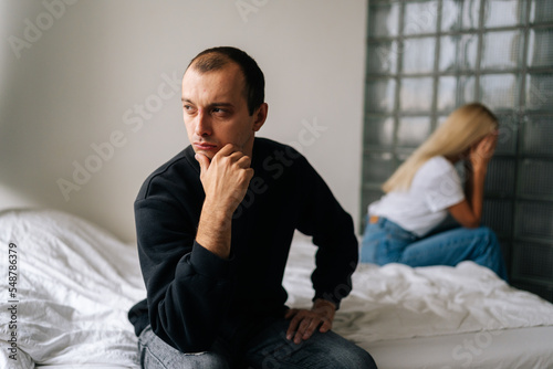 Portrait of serious young man pensive looking away not talking after dispute with sad wife sitting apart on bed. Depressed married couple sitting separate on couch having relationships problem.