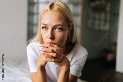 Close up face of thoughtful blonde female sitting alone in living room and serious looking at camera holding hands on chin  thinking over health problems  feeling sadness  boredom  apathy.