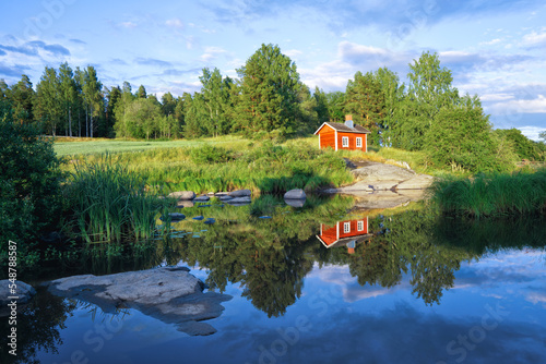 A small red cottage reflected in the river on a beautiful summer evening. An old traditional Finnish sauna cottage.