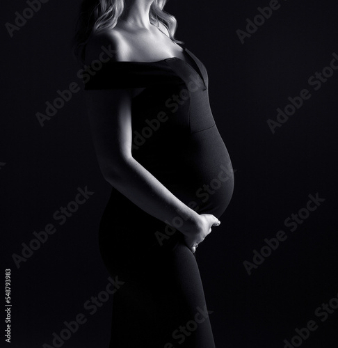 Pregnant woman in a black dress on a black background. Copy space. Pregnancy. Black and white. Woman in black.