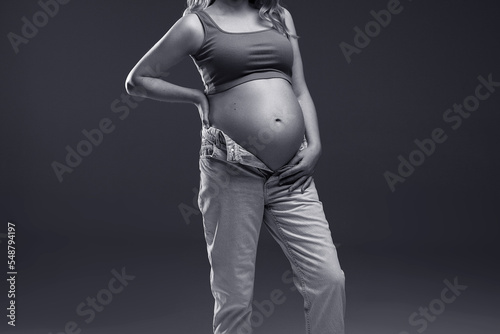 A pregnant woman in jeans stands on a gray background with a place for text. Studio shooting. Black and white photo. Pregnancy. Woman in blue jeans.