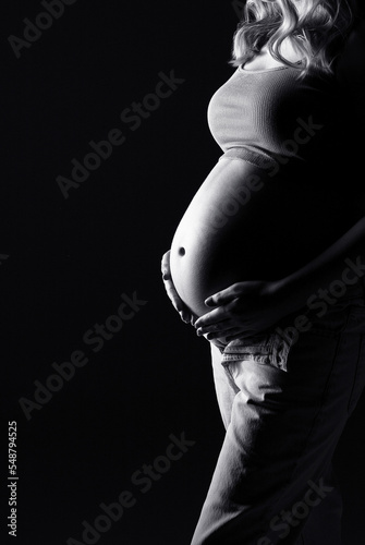 A pregnant woman in a jeans on a black background. A pregnant woman. Studio pregnancy photo shoot. Stylish pregnancy. Black and white photo. Model posing in studio.