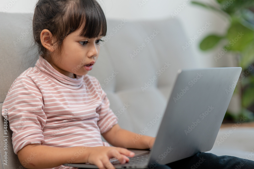 Asian Little school kid girl use laptop computer sitting on sofa alone at home. Child learning reading online social media content, play education lessons game chatting with friends. Asian Little school kid girl use laptop computer sitting on sofa alone at home. Child learning reading online social media content, play education lessons game chatting with friends.