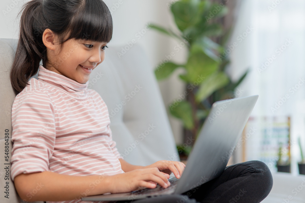 Asian Little school kid girl use laptop computer sitting on sofa alone at home. Child learning reading online social media content, play education lessons game chatting with friends. Asian Little school kid girl use laptop computer sitting on sofa alone at home. Child learning reading online social media content, play education lessons game chatting with friends.
