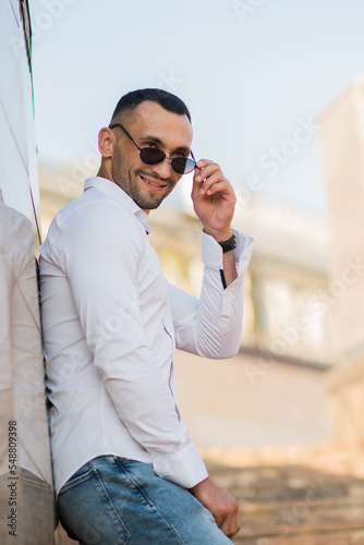 Handsome man in a trendy white shirt and sunglasses in the city. Handsome and charming man posing outdoors, urban style