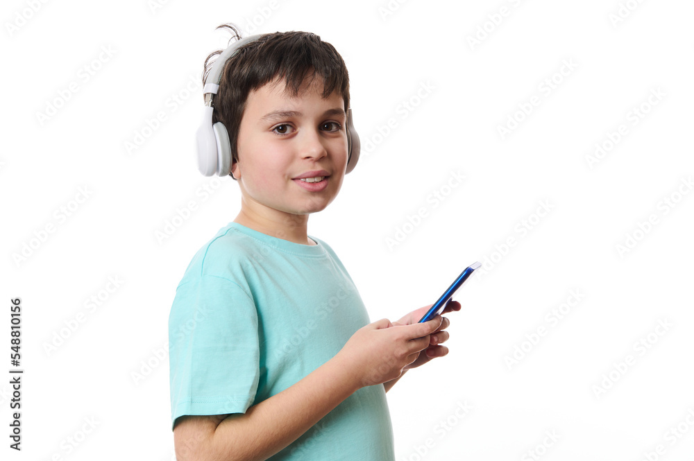 Isolated portrait on white background of a Caucasian smart teenage boy, wearing wireless headphones, using mobile phone, checking social media content, surfing the net, cutely smiles looking at camera Isolated portrait on white background of a Caucasian smart teenage boy, wearing wireless headphones, using mobile phone, checking social media content, surfing the net, cutely smiles looking at camera