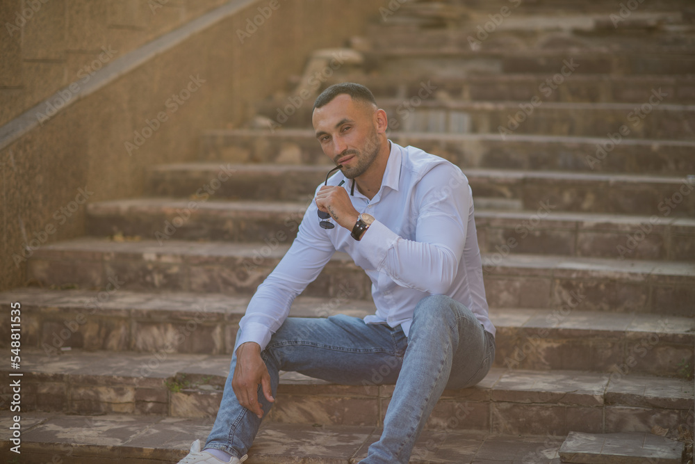 Attractive young man holding sunglasses sits on the steps in the city. stylish guy in a white shirt and jeans is resting in the city Attractive young man holding sunglasses sits on the steps in the city. stylish guy in a white shirt and jeans is resting in the city