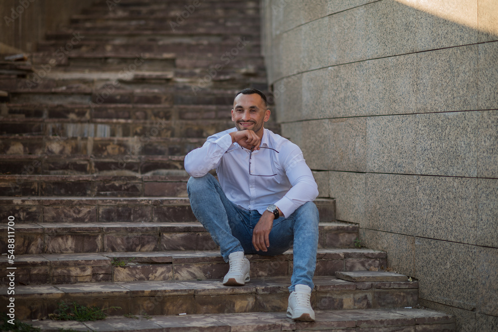Attractive young man holding sunglasses sits on the steps in the city. stylish guy in a white shirt and jeans is resting in the city Attractive young man holding sunglasses sits on the steps in the city. stylish guy in a white shirt and jeans is resting in the city
