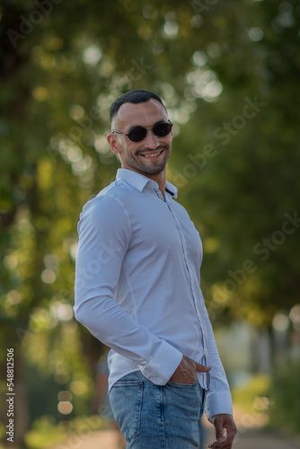 Attractive young man, wearing a white shirt and sunglasses, looks at the camera and smiles. a stylish guy in a white shirt and jeans walks in the city, a businessman at lunch