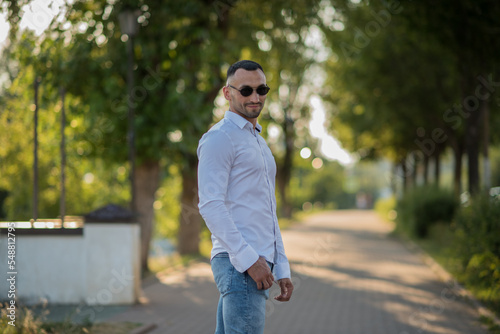 Attractive young man, wearing a white shirt and sunglasses, looks at the camera and smiles. a stylish guy in a white shirt and jeans walks in the city, a 
