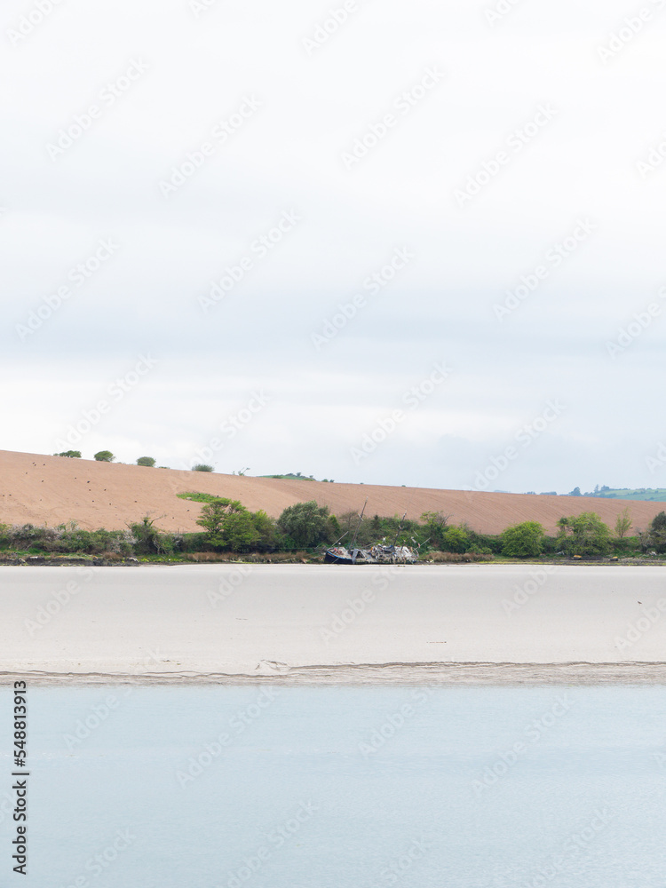 Sandy seashore. Calm water surface. The remains of an old ship on the shore. A plowed field. Minimalistic landscape.