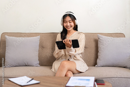 Female entrepreneur is sitting on big comfortable sofa and wearing headphone while holding tablet to watching