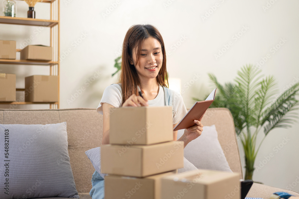 Young entrepreneur is reading customer data on notebook and writing address on cardboard boxes for home