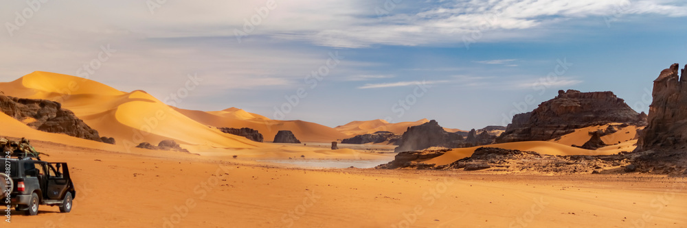 Panoramic view of Sahara Desert sand dune and rocky mountain off road nature.Tadrart Rouge, Djanet, Illizi. Erg dusty road. Orange colored sandstones. A 4X4 car loading wood parked with door opened.