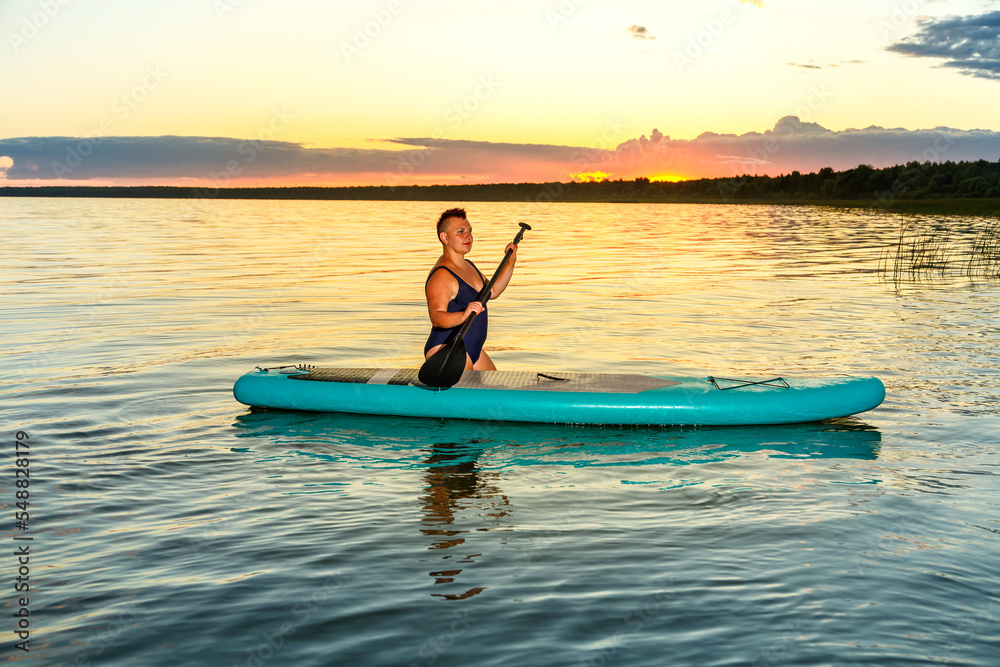 a woman in a bathing suit with a mohawk sits on a sup board at sunset in the water.