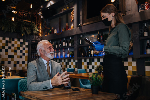 Waitress serves and takes the order from the senior businessman at the restaurant. She wears a protective mask as part of security measures against the Coronavirus pandemic.