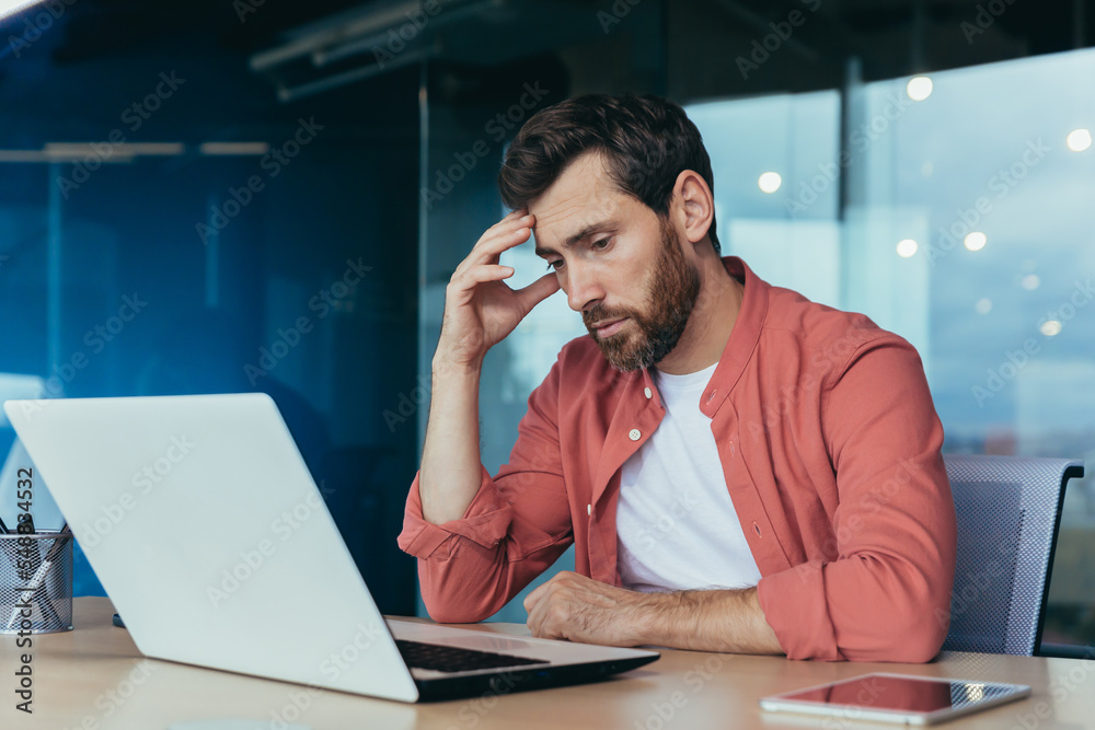Sad and thinking programmer in red shirt working on laptop inside office, mature man with beard. Sad and thinking programmer in red shirt working on laptop inside office, mature man with beard.