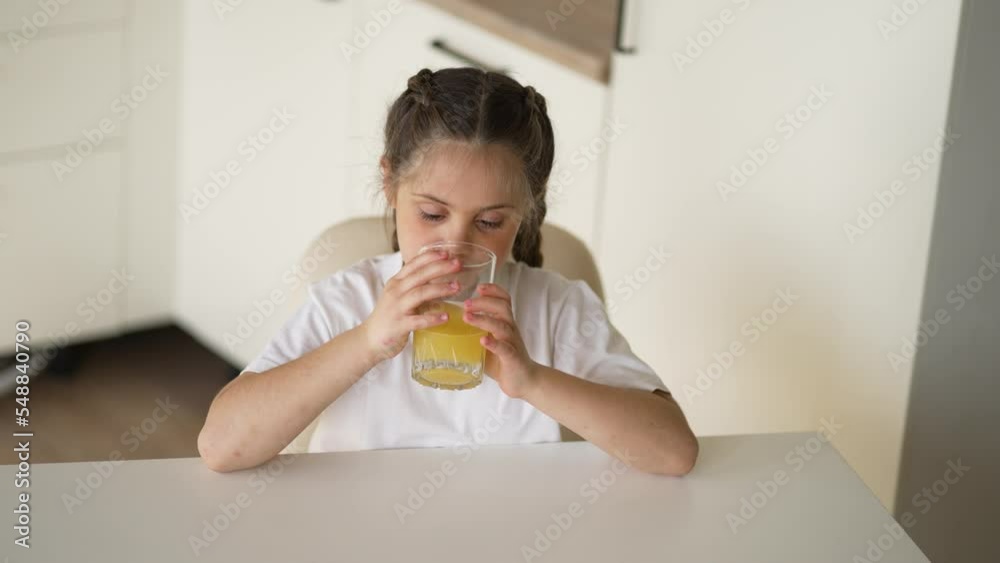 Girl sits at an empty table with a glass of orange juice. Healthy vitamin baby food. Kid with fruit drink in the kitchen. Funny kid drinks juice. Girl healthy food. Child eating breakfast in kitchen Girl sits at an empty table with a glass of orange juice. Healthy vitamin baby food. Kid with fruit drink in the kitchen. Funny kid drinks juice. Girl healthy food. Child eating breakfast in kitchen