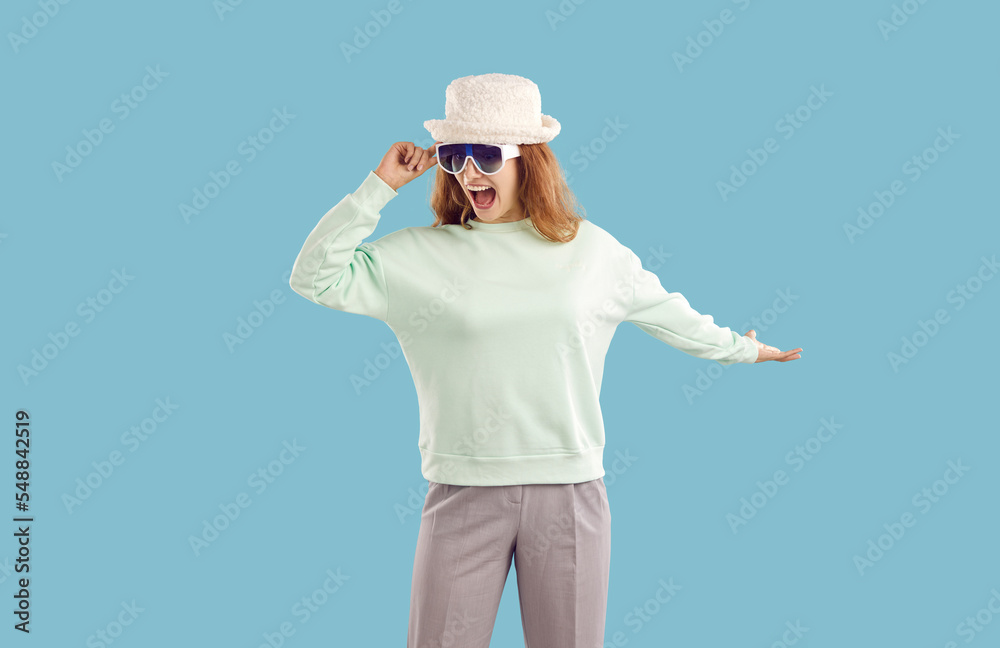 Impressed happy funny cheerful young blonde woman in wool furry bucket hat, sunglasses, mint cotton sweatshirt, grey trousers is dancing with arm stretched out to the side on blue background. Impressed happy funny cheerful young blonde woman in wool furry bucket hat, sunglasses, mint cotton sweatshirt, grey trousers is dancing with arm stretched out to the side on blue background.