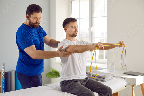 Physiotherapy specialist and his male patient use special equipment during rehabilitation. Young man doing remedial physical exercises with resistance band under supervision of serious physiotherapist