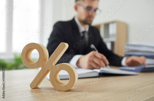 Close up of wooden percent sign on table as symbol of corporate tax and interest rate. Percent sign standing on background of serious busy man in suit calculating on calculator. Blurred background. photo