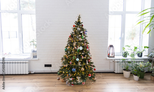 Christmas tree in the white interior of a house with large windows. Glowing fairy lights garlands interior decoration of the studio room. Potted plants in the home