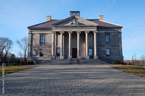 The exterior facade of a historic government building with large vintage marble pillars or columns  red door  blue sky  trees  and a brick entrance leading up steps to the large antique stone building