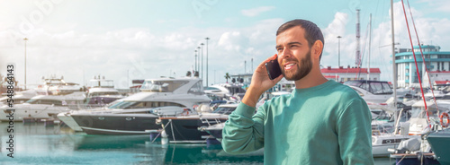 Tablou pe pânză Caucasian male in green pullover talking on phone port against backdrop of expensive yachts