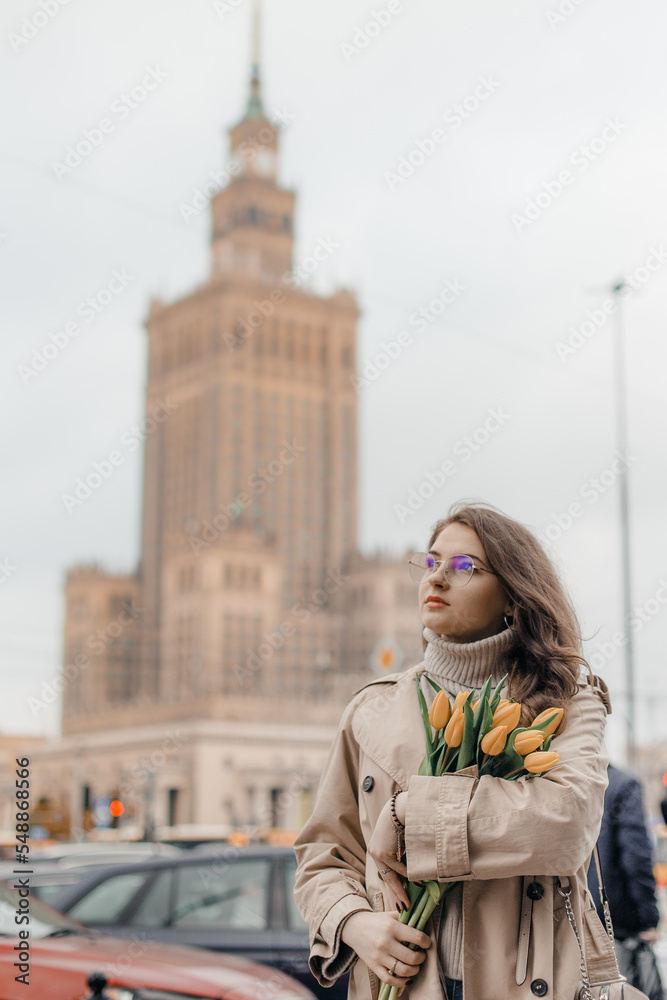 Fototapeta premium A young girl in glasses with flowers against the background of the Palace of Culture in Warsaw. A pretty girl in glasses with flowers walks in Warsaw.